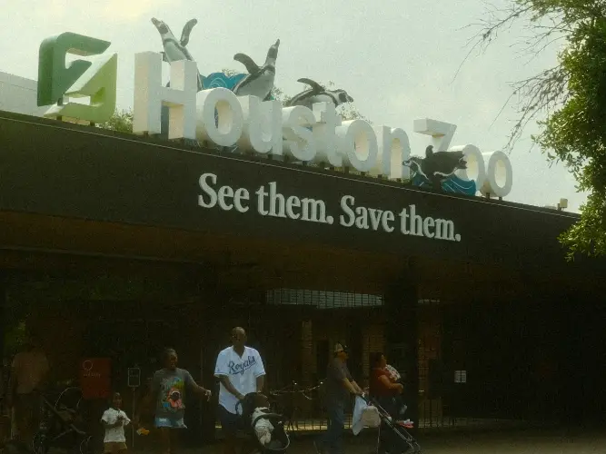 The exterior of the Houston Zoo entrance with people walking in front of it.